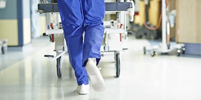 Los pies de un proveedor de atención médica alejándose con ruedas de camilla en el fondo.
