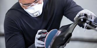 Trabajador usando guantes de protección operando una lijadora con un disco abrasivo azul sobre la superficie de un vehículo.
