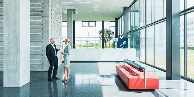 Plano amplio del vestíbulo de un moderno edificio de oficinas rodeado de paredes con ventanas y dos empresarios hablando.
