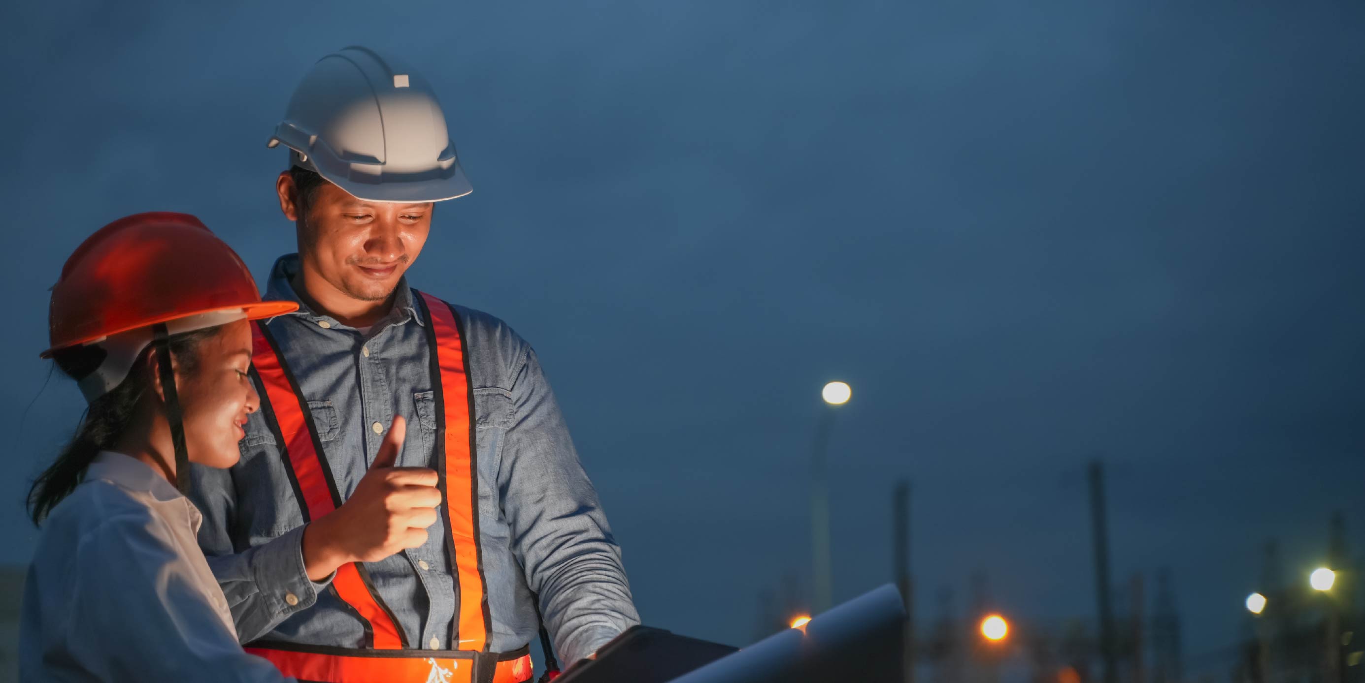 Personal de obra con cascos y chalecos reflectantes revisa planos al anochecer. 
