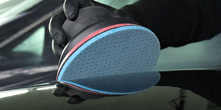 Una persona con guantes negros sostiene una lijadora eléctrica con un disco abrasivo flexible de espuma 3M™ Hookit™ azul, colocada sobre una superficie brillante, lista para lijar.
