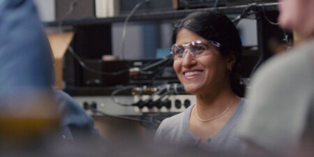A woman smiling, wearing safety glasses in lab with electronic equipment behind her.
