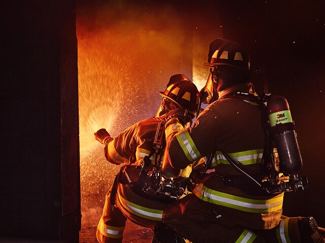 Bomberos con equipo completo combaten un fuego intenso dentro de una estructura, iluminados por chispas y llamas mientras avanzan con precaución.