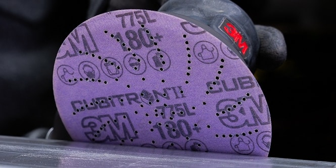 Un disco de lijado 3M Cubitron II 775L de color morado, unido a una lijadora eléctrica, que muestra su granulometría, patrón de perforación y logotipo impreso mientras se utiliza sobre una superficie.
