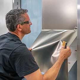 Hombre instalando acabado metálico plateado 3M DINOC en la puerta del elevador.