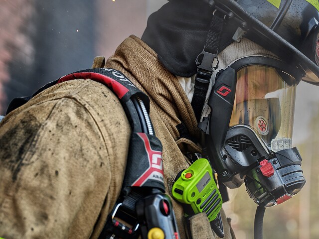 Bombero en primer plano con máscara de respiración y equipo de protección, sosteniendo un dispositivo de comunicación en un entorno con humo.