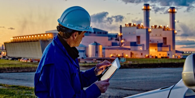 Trabajador con casco blanco y uniforme azul mirando la pantalla de un dispositivo electrónico.
