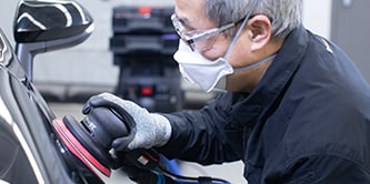 Trabajador usando gafas de seguridad, guantes y mascarilla protectora mientras utiliza una pulidora  para preparar la superficie de un vehículo.
