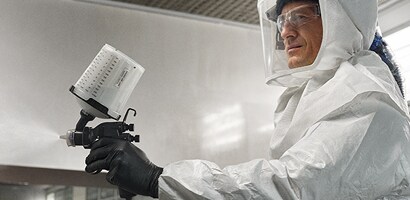 Un trabajador con equipo de protección completo, incluyendo traje y guantes, utiliza una pistola de pintura en un entorno controlado.
