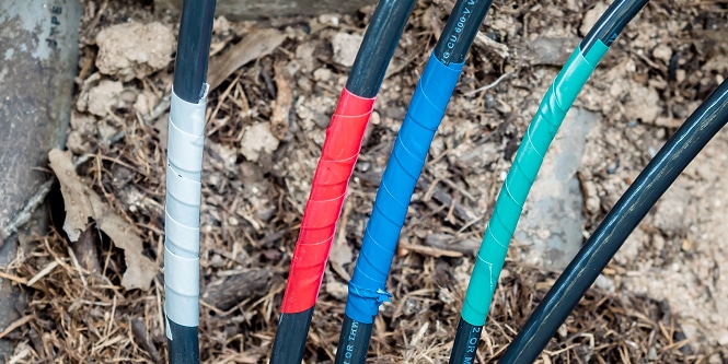 Cuatro cables exteriores envueltos con cinta de colores - plateado, rojo, azul y verde - que salen del suelo en una zona de tierra y mantillo.
