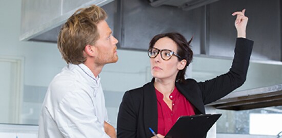 In a commercial kitchen, a woman with a clipboard points up as a chef looks on.
