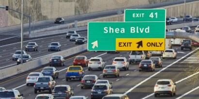 A large road sign is visible from a high angle above a busy highway, directing rush-hour drivers to an exit.
