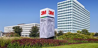 Imagen del exterior la empresa 3M. Se encuentran 2 edificios, plantas y árboles rodeándolos.
