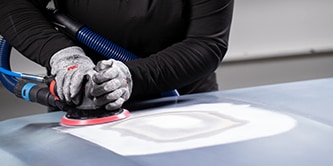 Trabajador usando guantes de protección utilizando una lijadora orbital con un disco de abrasivo para alisar una sección preparada de la superficie de un vehículo.
