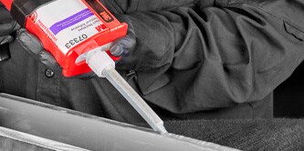 Técnico aplicando masilla de reparación automotriz 3M sobre una superficie metálica utilizando una espátula, con el envase del producto visible en primer plano.
