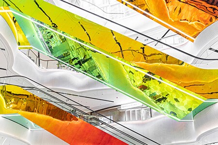 Centro comercial de varios niveles con paredes blancas y escaleras mecánicas suspendidas cubiertas con acabados reflectantes y coloridos de vidrio dicroico 3M en tonos de rojo, amarillo, naranja y verde.
