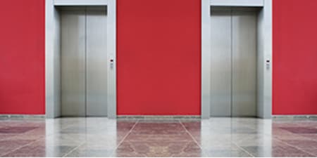 Imagen de las puertas de dos ascensores con puertas cerradas. Paredes rojas y piso gris brillante.
