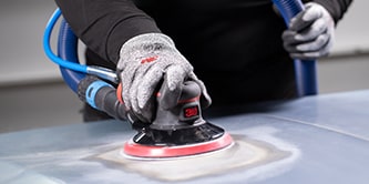 Trabajador usando guantes de protección utilizando una lijadora orbital de 3M con un disco abrasivo para alisar la superficie de un vehículo.

