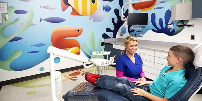 Un niño en la silla del dentista y un técnico dental sonriente en el consultorio del dentista, con una vibrante ilustración del océano impresa en películas gráficas de 3M.

