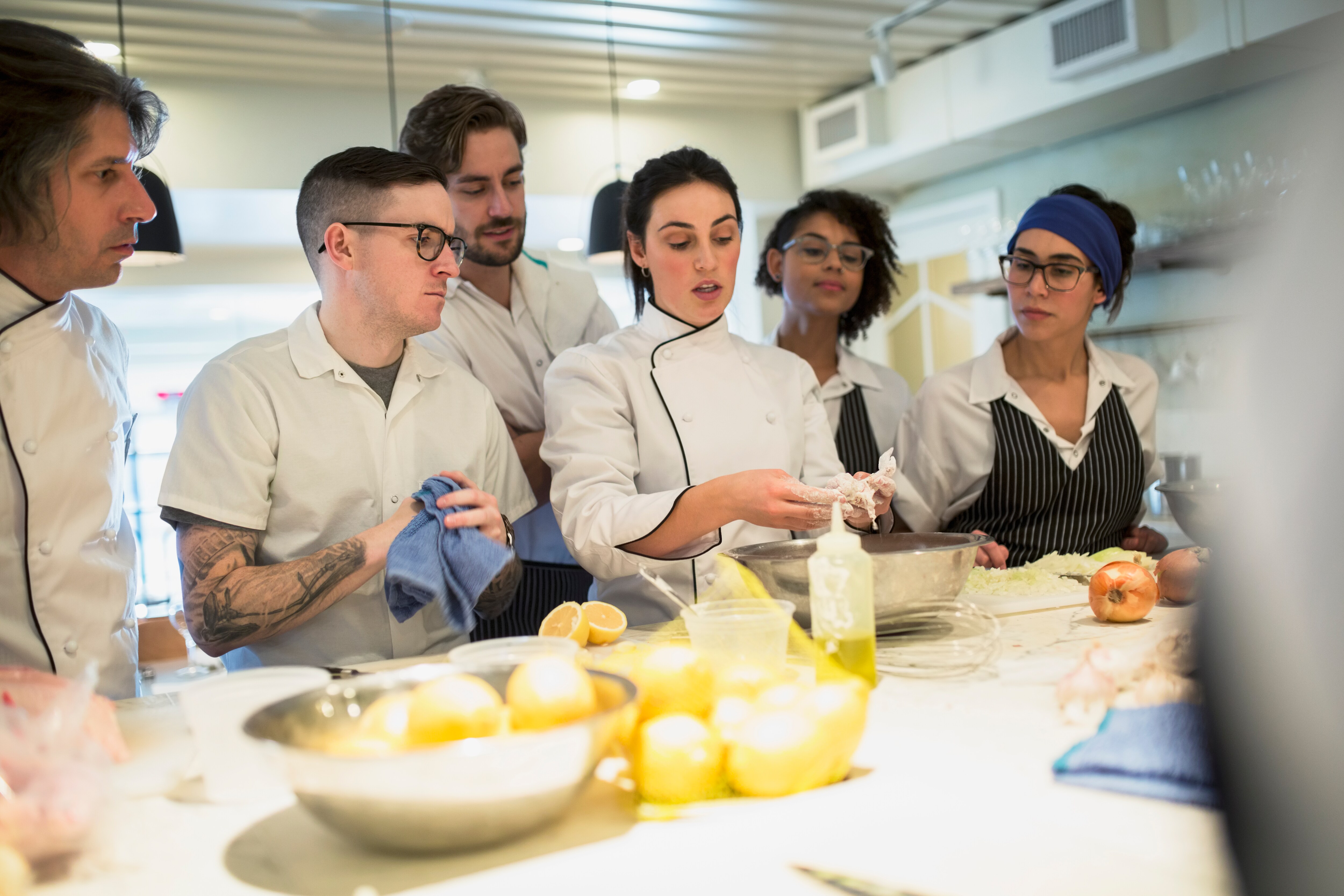 Four onlooking commercial kitchen workers surround a chef demonstrating a food preparation technique.
