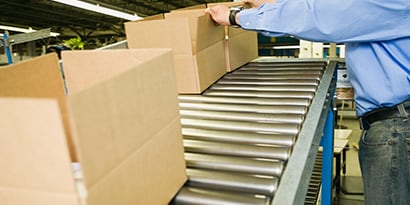 Imagen de cajas abiertas moviéndose a lo largo de una cinta transportadora hacia un trabajador que dobla las solapas para preparar el cierre.
