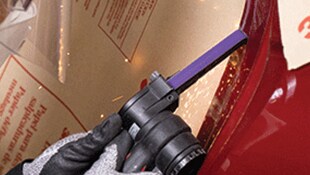 Una mano con un guante que utiliza una herramienta eléctrica manual con una banda esmeriladora estrecha sobre una superficie metálica y que produce pequeñas chispas
