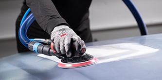 Trabajador usando guantes de protección operando una lijadora orbital 3M con un disco abrasivo para alisar la superficie de un vehículo.
