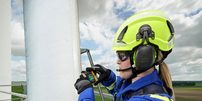 sideview of woman on wind tower wearing climbing helmet and earmuffs
