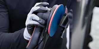 Trabajador usando guantes de protección sosteniendo una lijadora con un disco abrasivo azul mientras se prepara para lijar la superficie de un vehículo.
