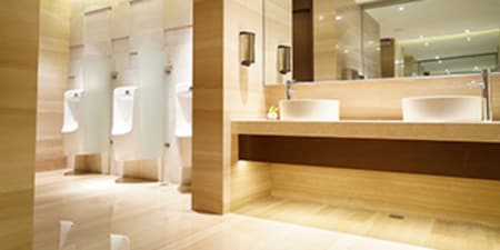 Imagen de un baño con tres urinales separados por mamparas. Dos lavaderos de manos y un espejo rectangular. Espacios luminosos.

