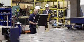 Video still of workers at a machine on the assembly line
