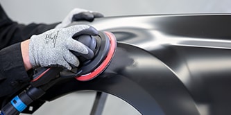 Trabajador usando guantes de protección utilizando una lijadora orbital con un disco abrasivo para alisar un panel negro de vehículo.
