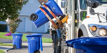 Un contenedor de reciclaje azul siendo levantado por un camión de reciclaje.
